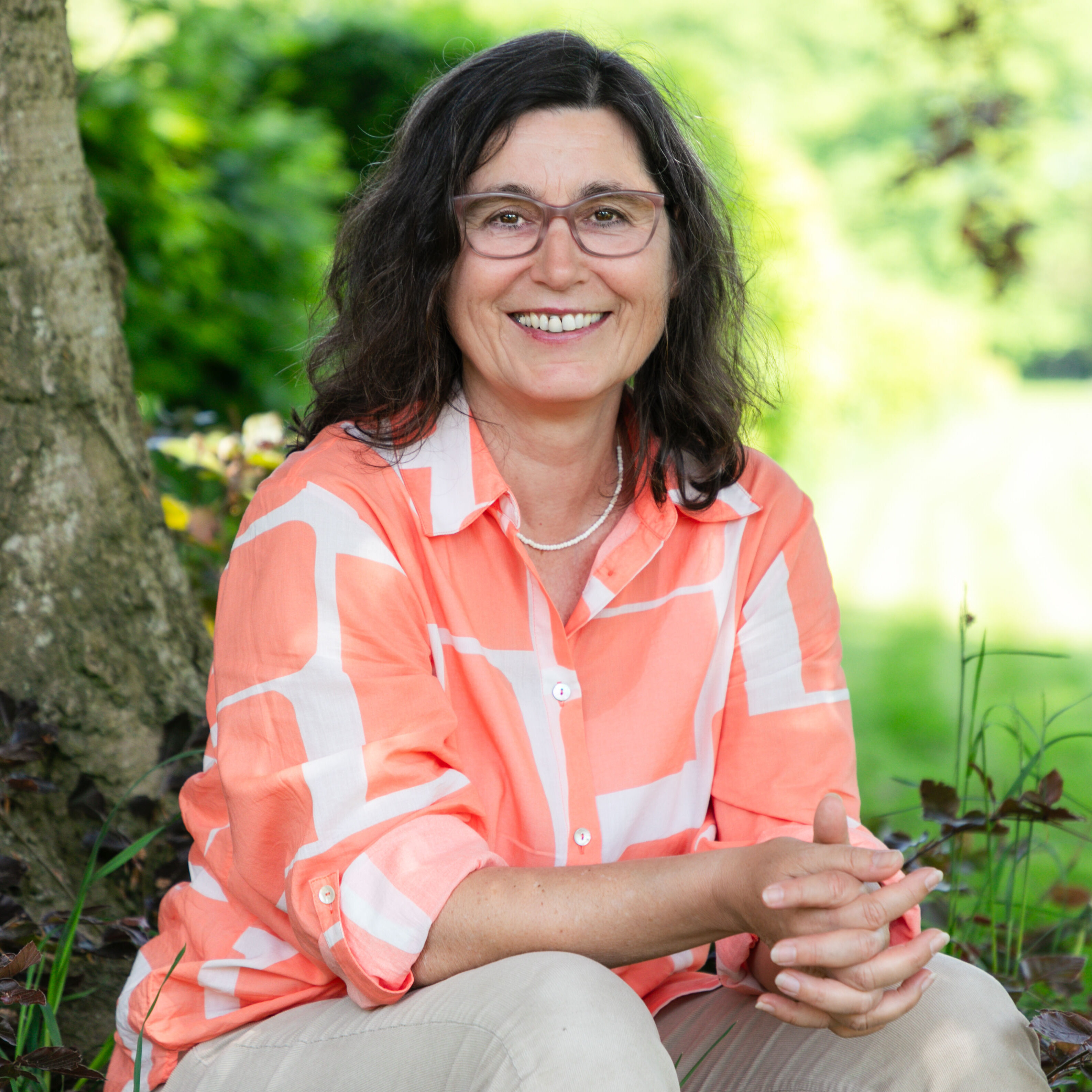 Dozentin Monika Weber. Lächelnd mit weiß-orangener Bluse vor einem Baum sitzend.