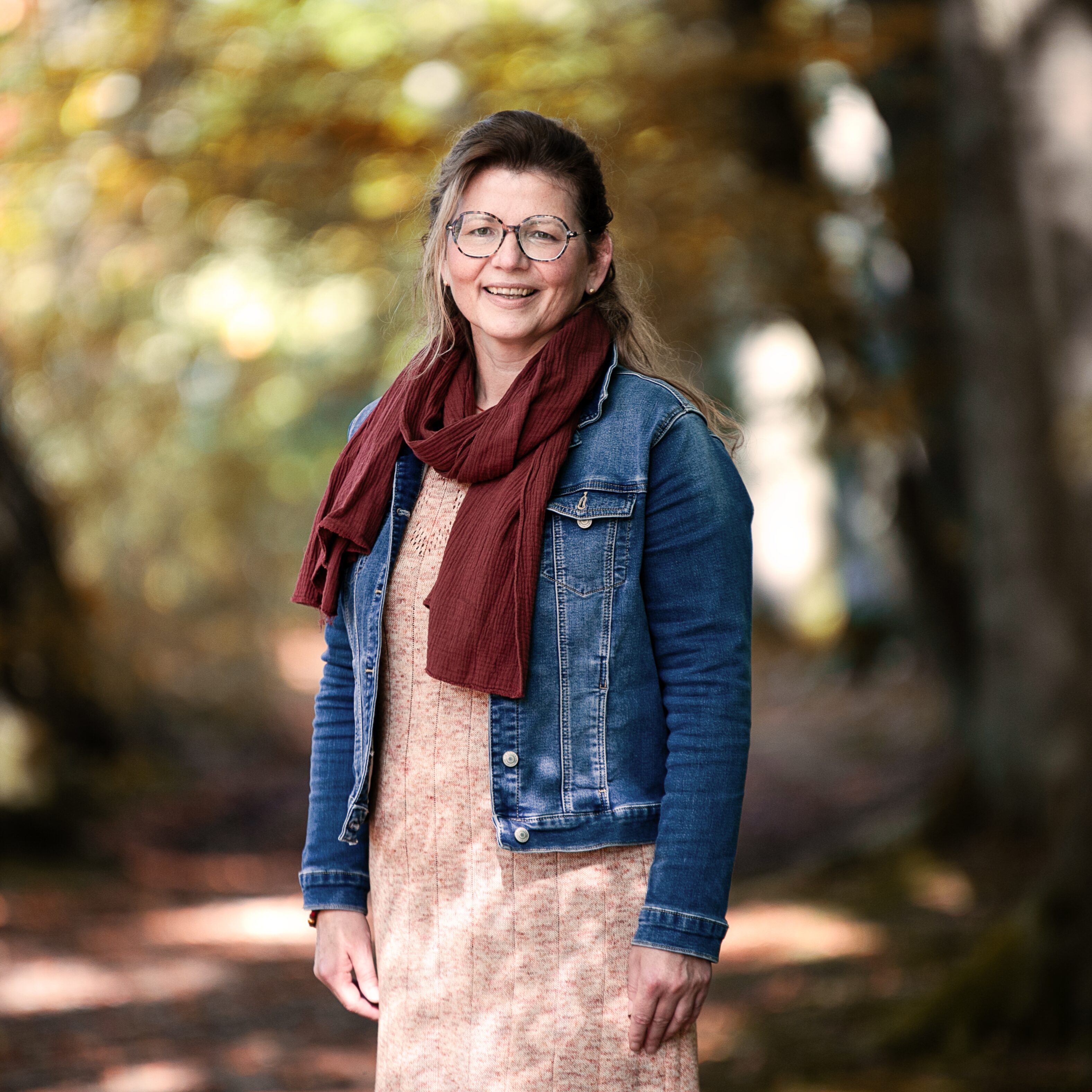 Foto der Dozentin Anke Brehl mit Jeansjacke, braunmeliertem Kleid, rostrotem Schal und Brille. Lächelnd.
