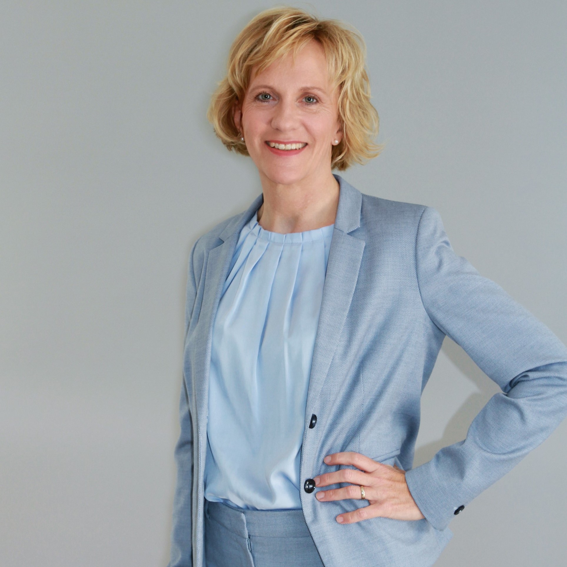 Dozentin Birgit Happel. Frau in Helblauen Anzug. Eine Hand in die Hüfte gestützt. Blonde, kinlange Haare, lächelnd.