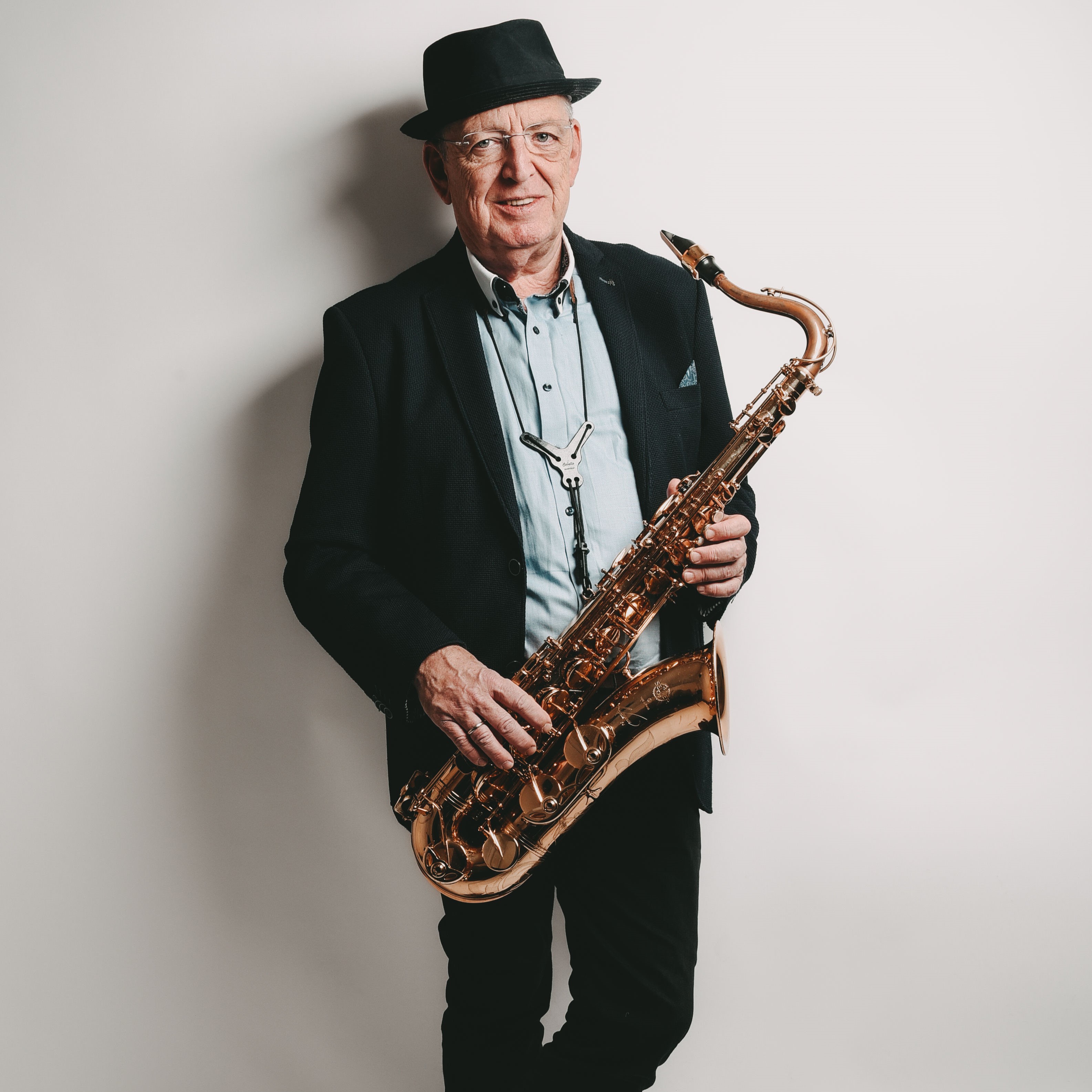 Dozent Dirk Zygar. Ein älterer Herr in dunklem Anzug, hellem Hemd, und dunklem Hut hält ein Saxophone in beiden Händen. Er steht vor einer weißen Wand.