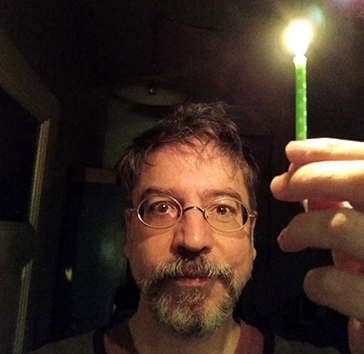 Dozent Matthias Vogel. Mann mit Brille und Bart in Frontansicht. Er hält eine Kerze ins Foto.