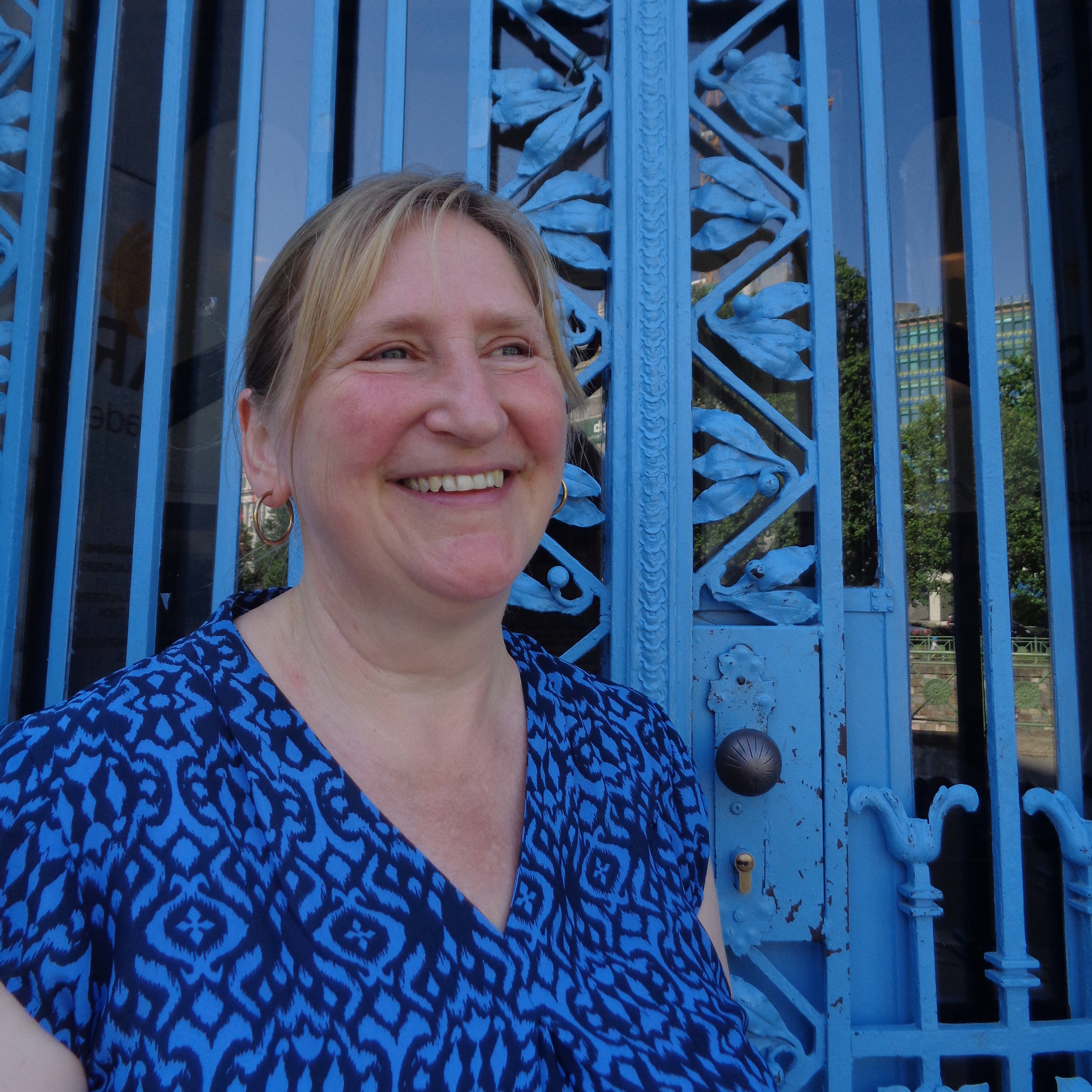 Dozentin Gloria Thom. Lächelnd mit blauer Bluse vor Blauer Tür mit Ornamenten.