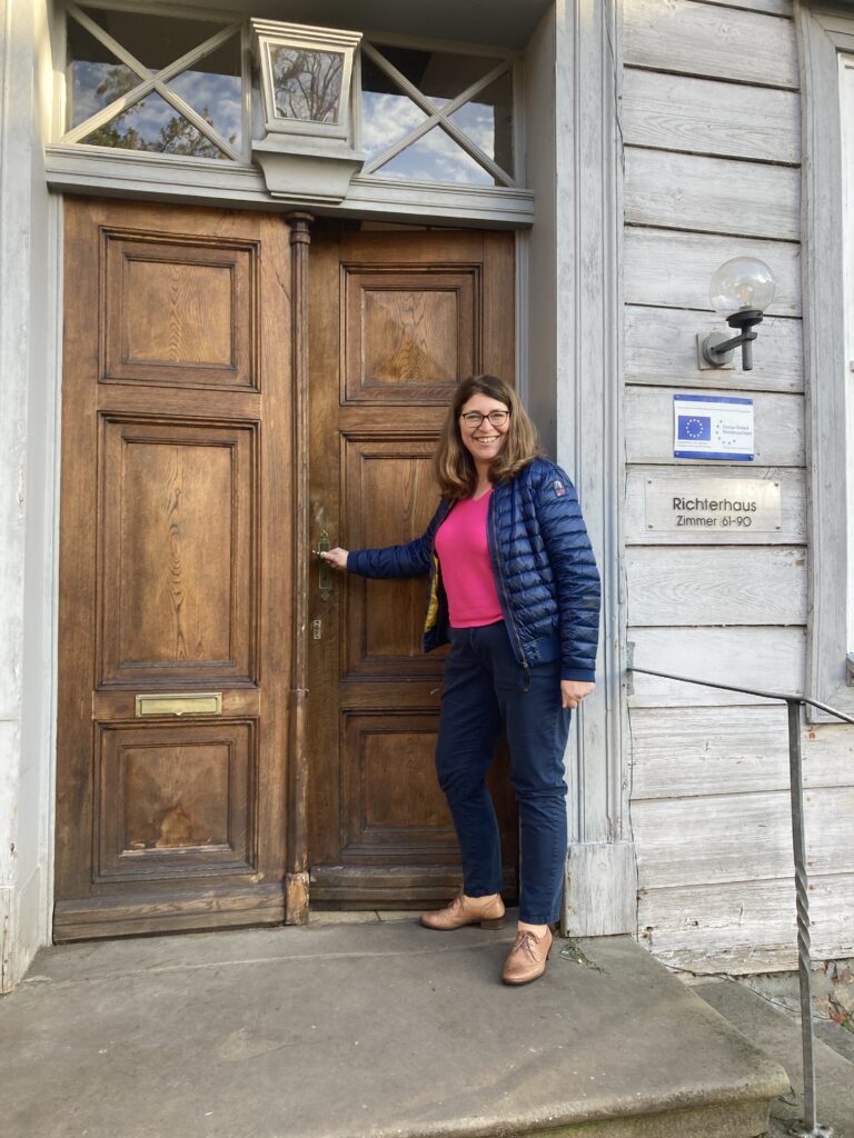 Frau Anke Matthes steht vor einer großen braunen Tür, um diese zu öffnen. Dabei blickt sie in die Kamera und lächelt.